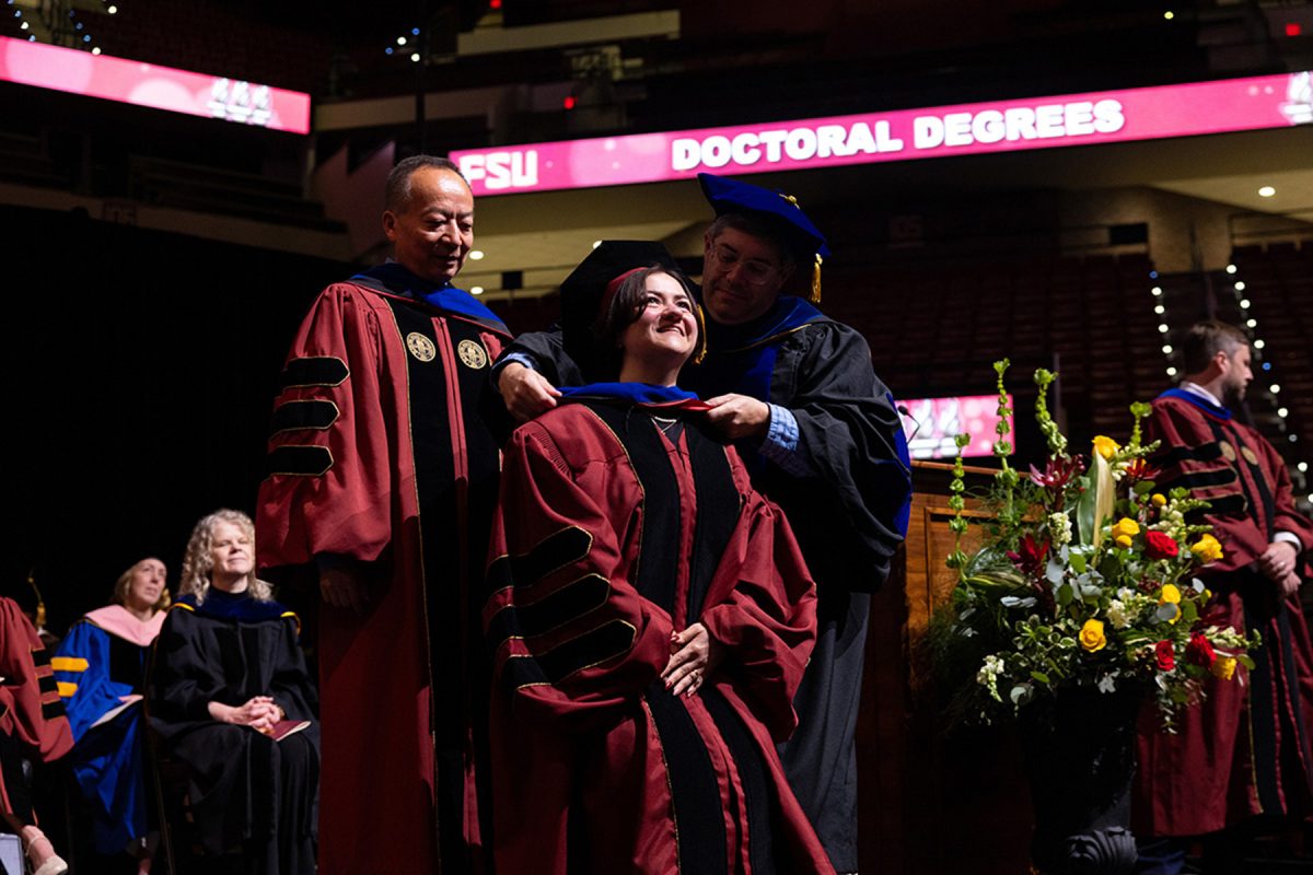 A female doctoral candidate beams as two faculty members assist in placing her academic hood over her shoulders during a commencement ceremony.