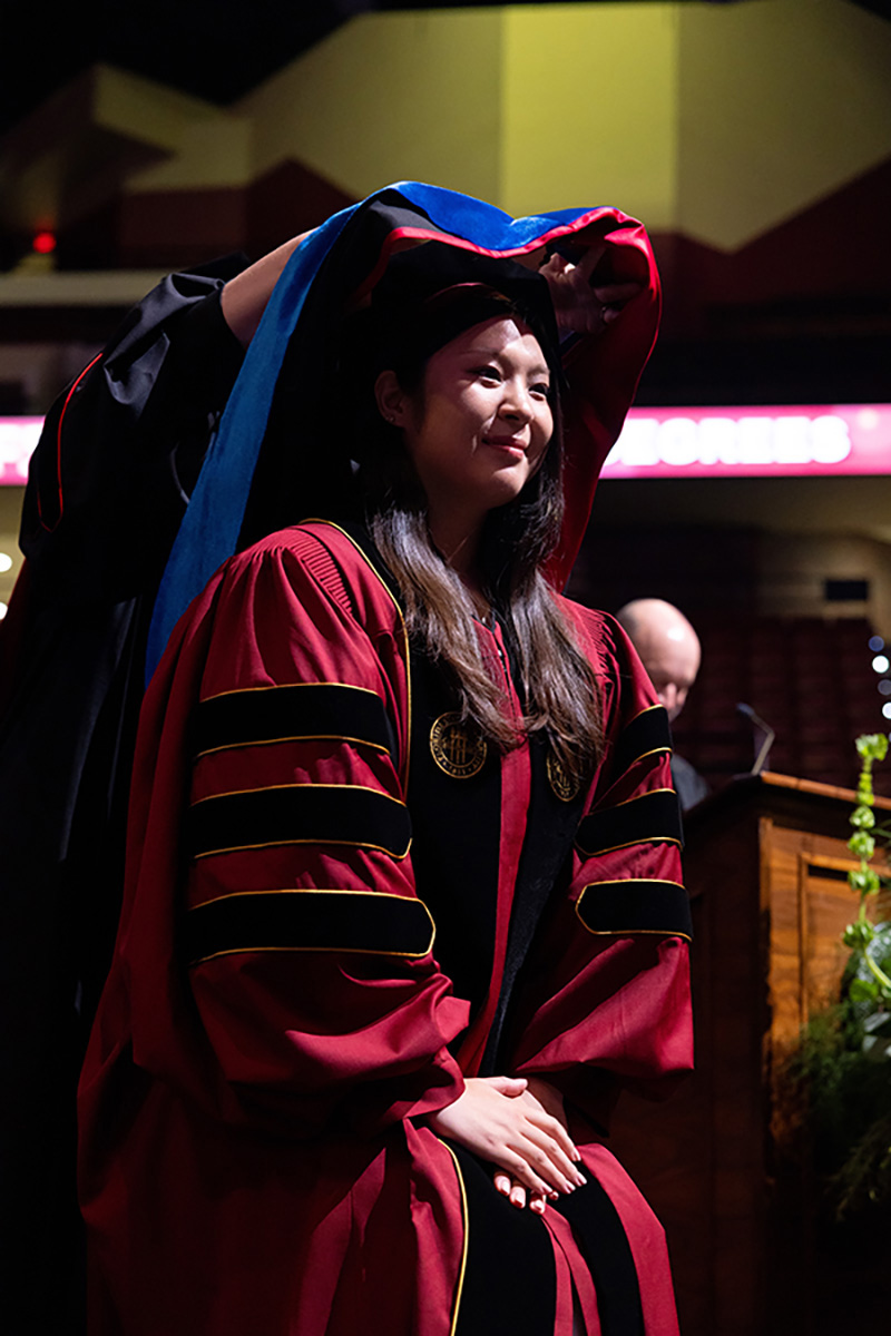 A graduating woman sits serenely with her hands folded as a blue and red velvet hood is draped around her head during a doctoral ceremony.
