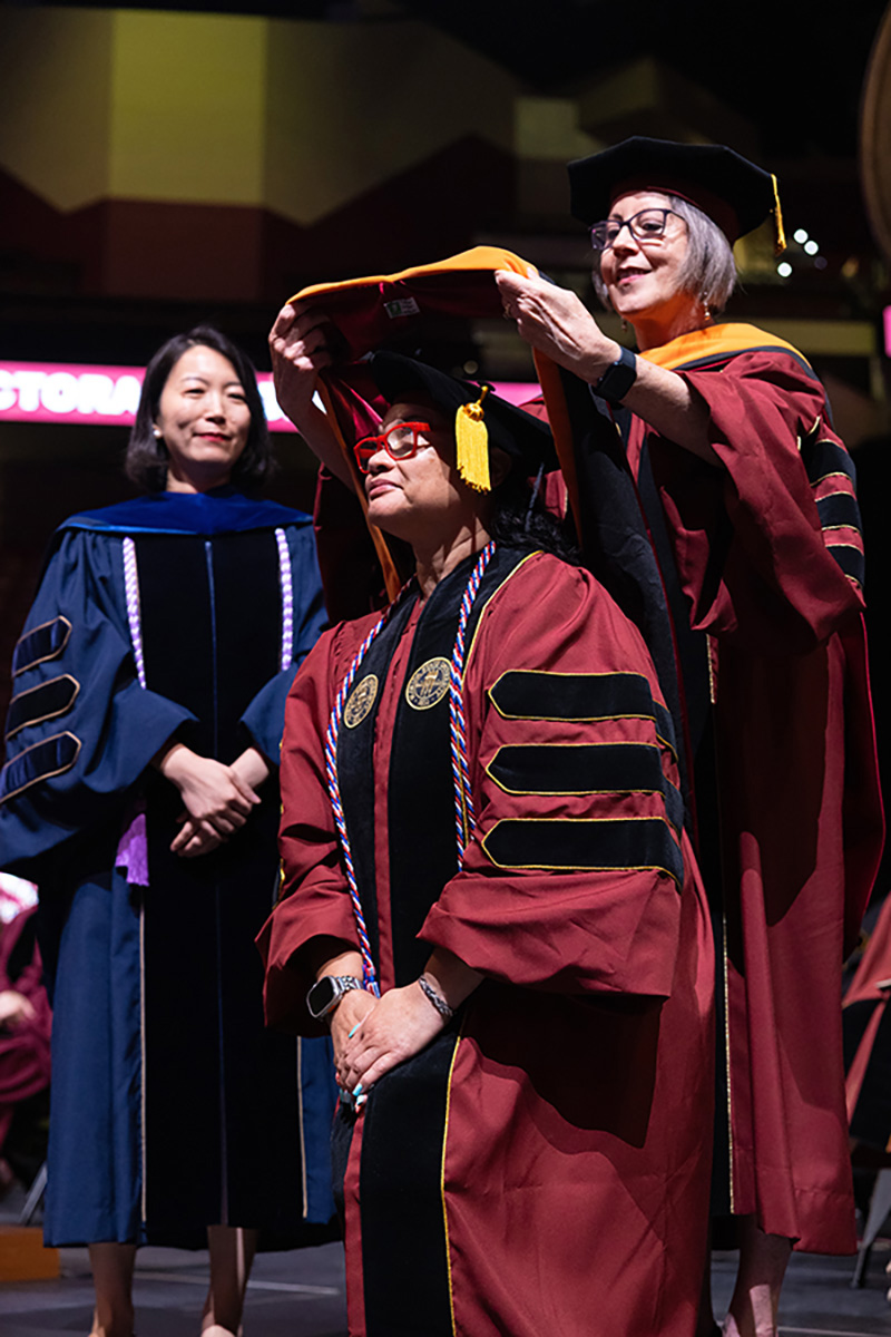 A graduate wearing red-framed glasses receives her doctoral hood from a faculty member during a commencement ceremony.