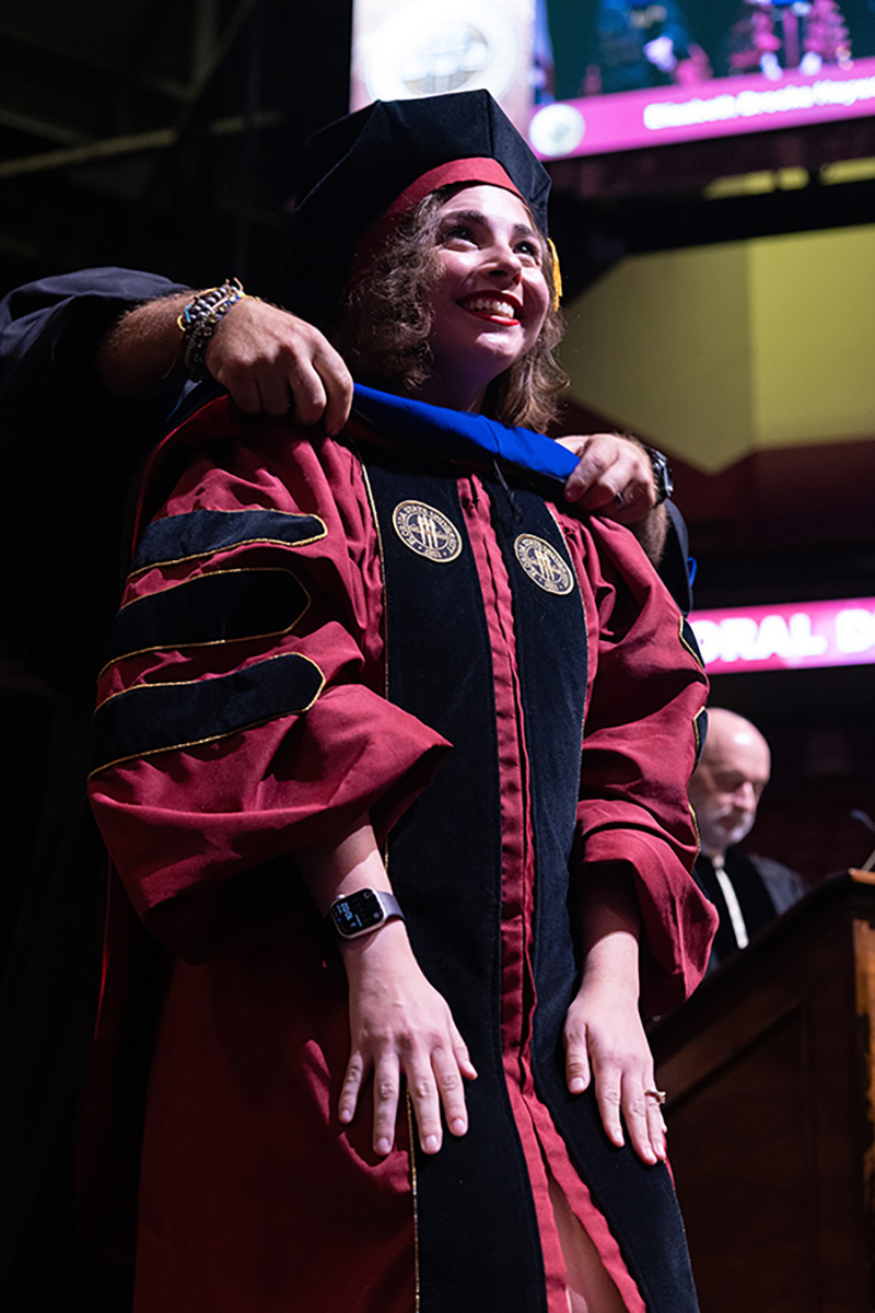 A low-angle shot of a joyful doctoral graduate smiling upward as an official arranges her academic hood.