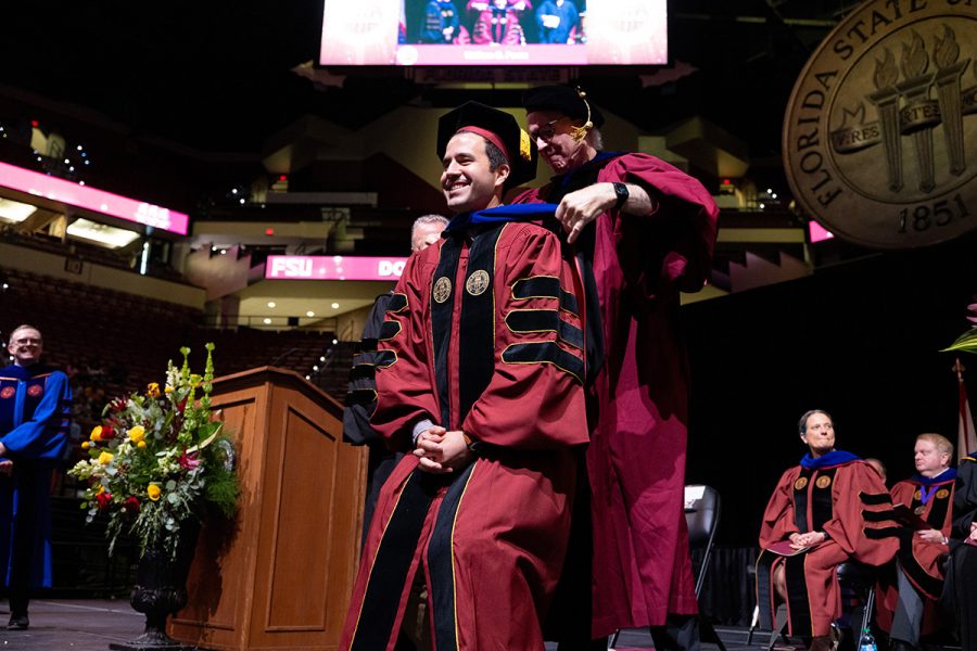 A smiling graduate in doctoral regalia is hooded by a university official on stage during a ceremony.