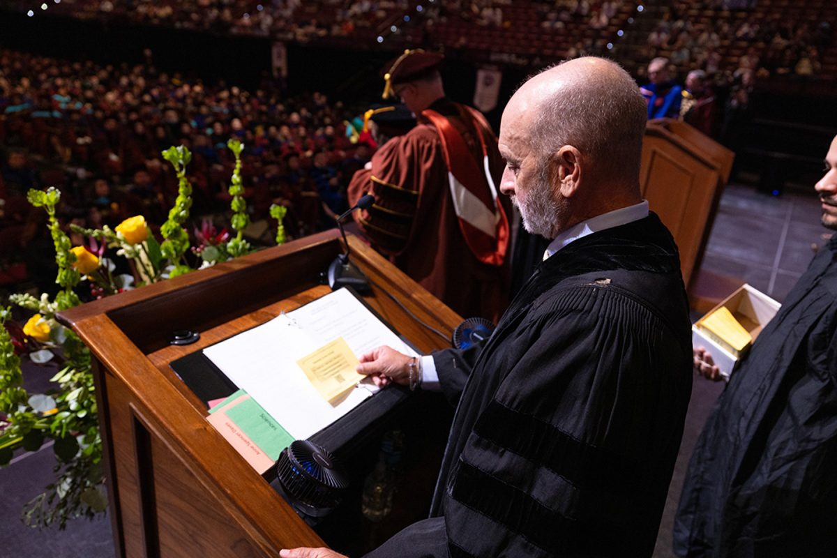 A speaker in black regalia reads from notes at a podium overlooking a crowd during a graduation ceremony.