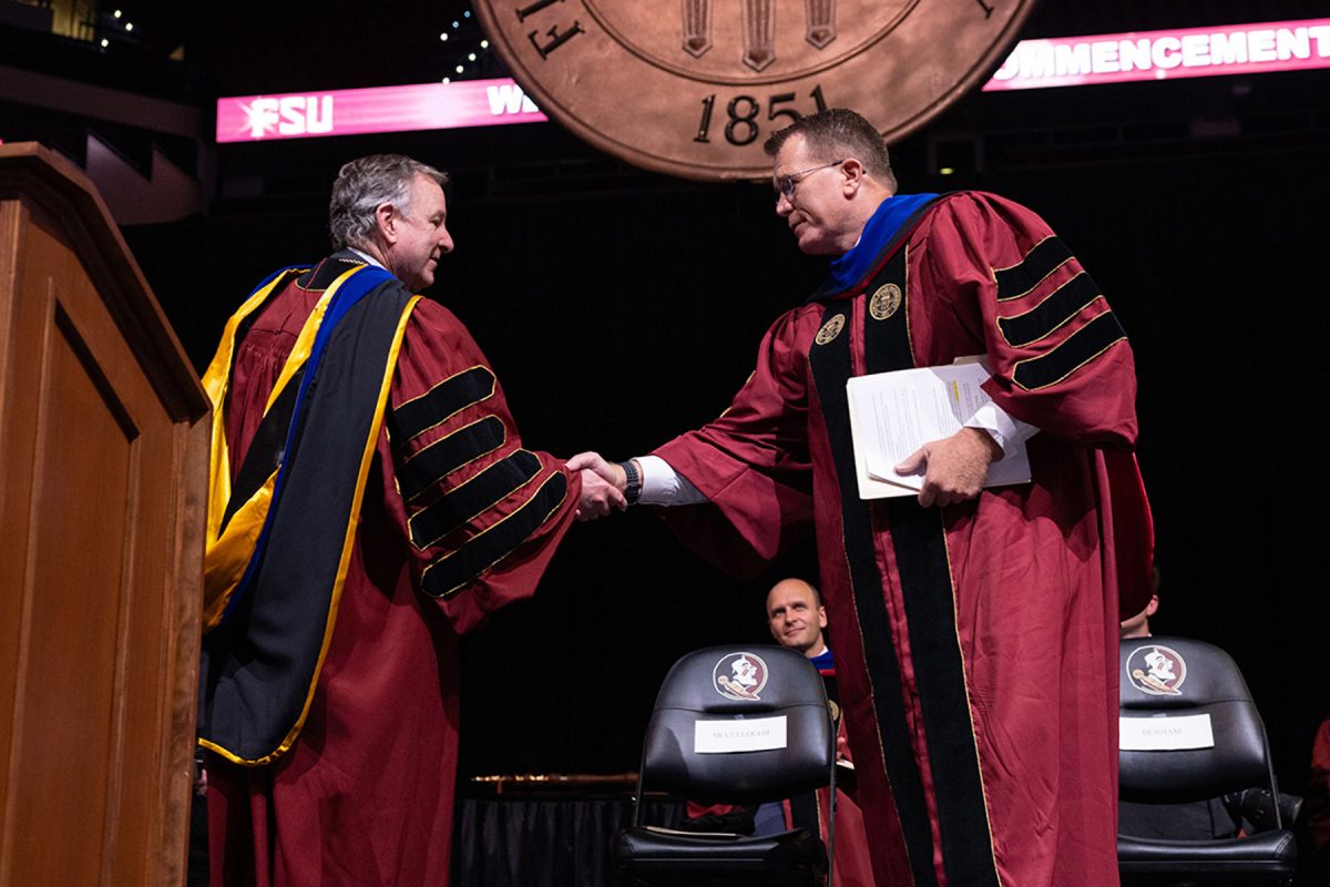 FSU President Richard McCullough shakes hands with another academic official wearing regalia on stage under the large university seal.