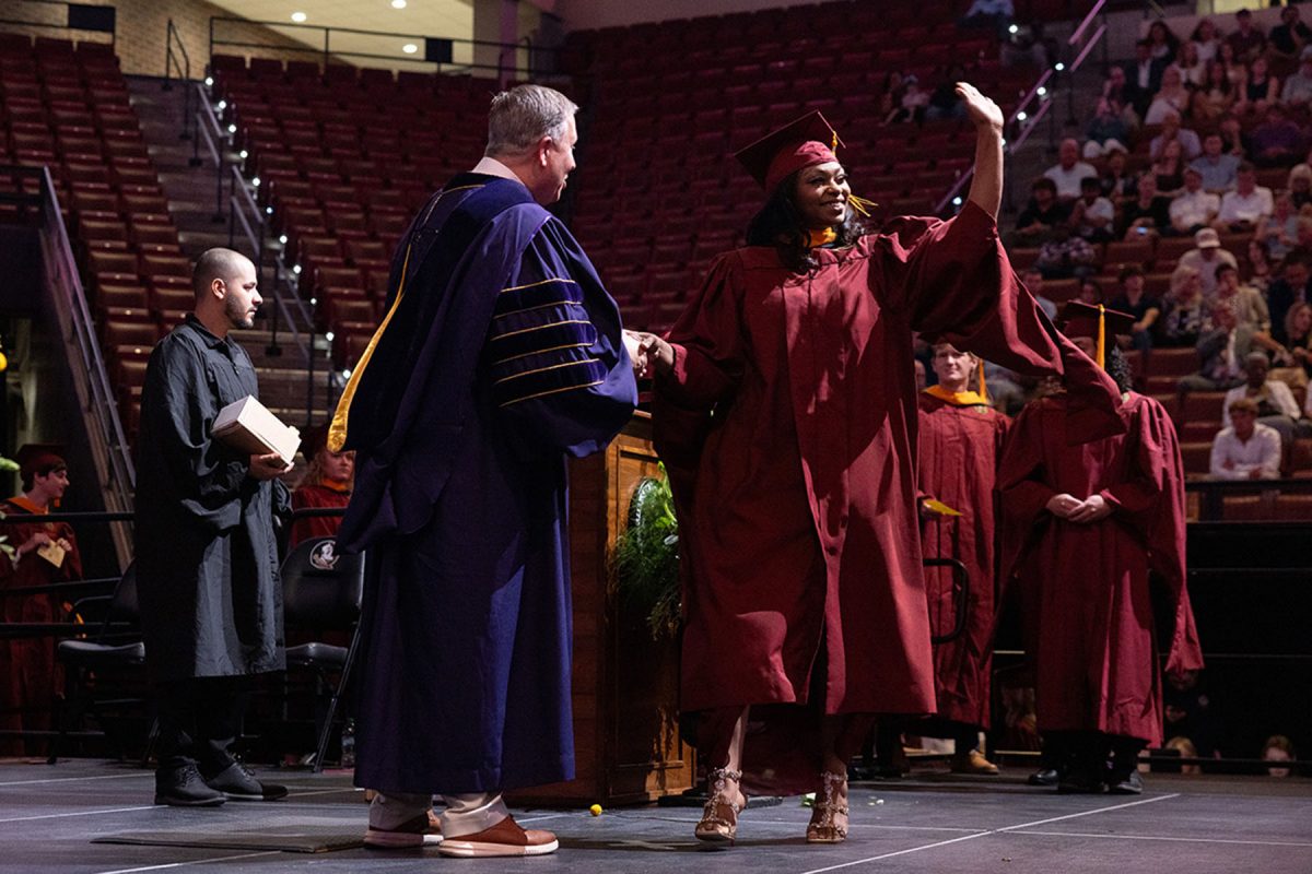 A smiling graduate waves high to the crowd while shaking hands with a faculty member on stage.