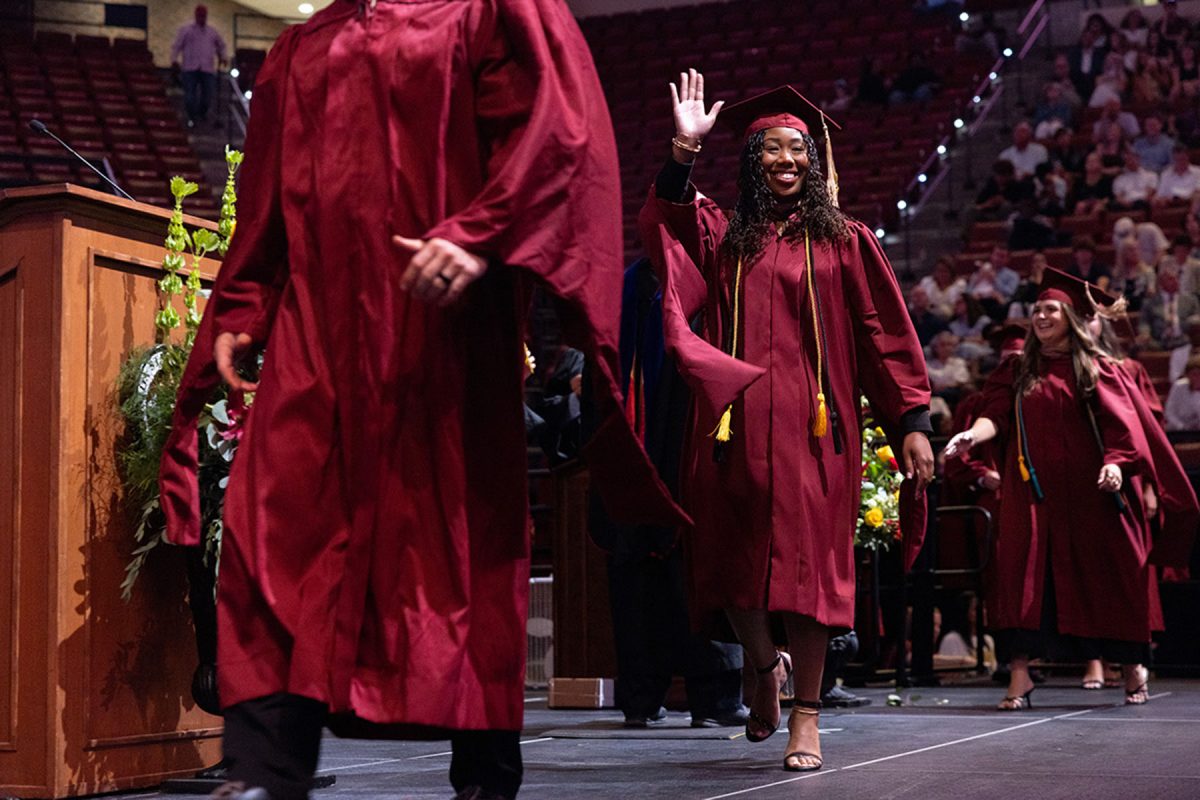 A smiling graduate in a maroon gown waves to the audience as she walks across the stage behind another graduate.