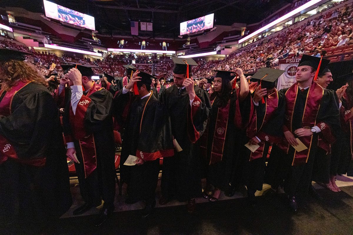 A wide view shows a row of graduates standing together in an arena, adjusting their mortarboards and caps during the ceremony.