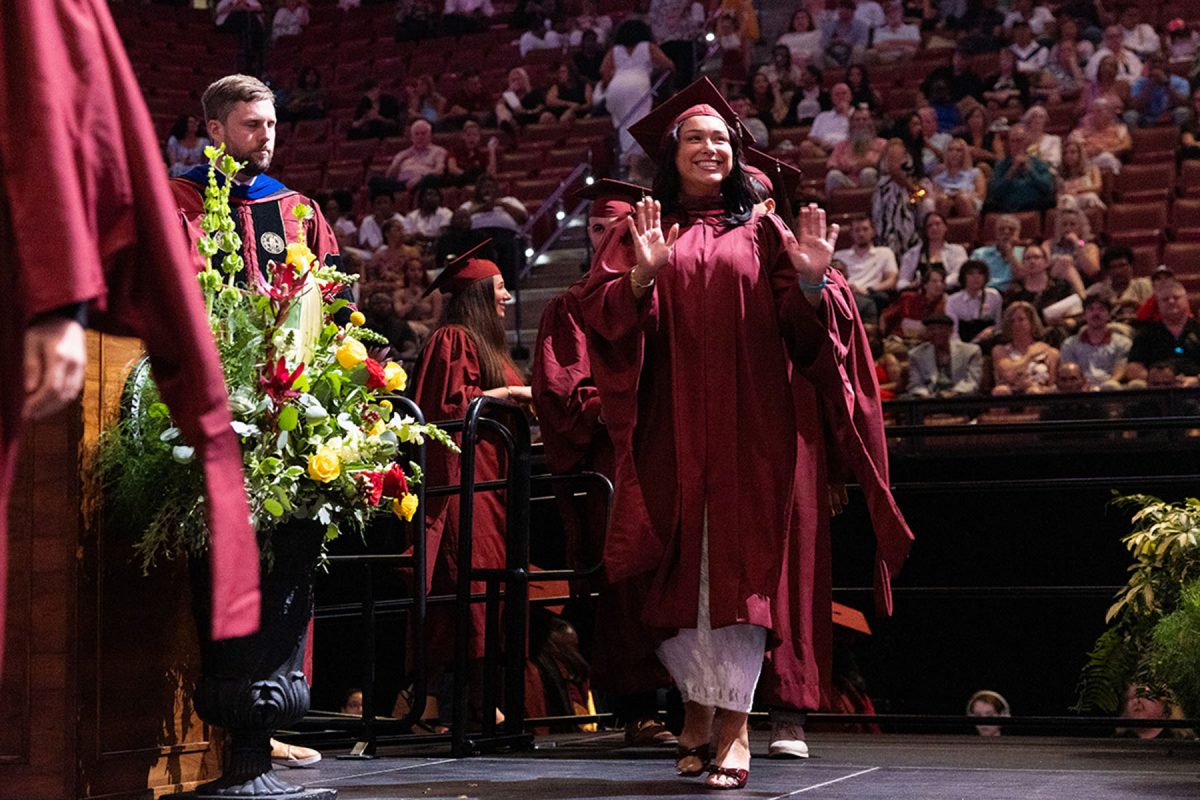 A smiling graduate in a maroon gown raises both hands to wave to the audience as she walks across the stage.