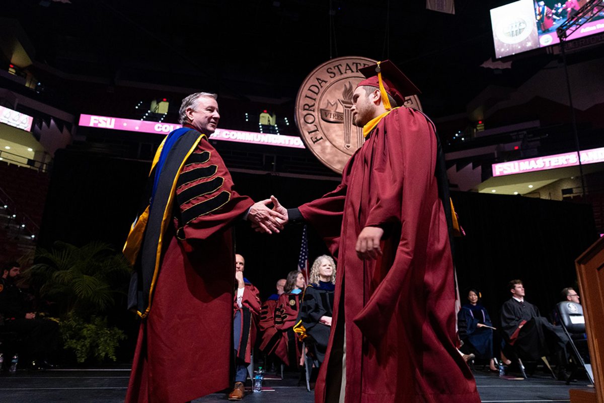 FSU President Richard McCullough shakes hands with a male graduate wearing a maroon cap and gown in front of the Florida State University seal.