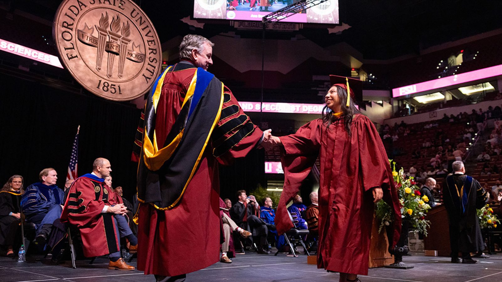 FSU President Richard McCullough shakes hands with a smiling graduate on stage beneath the large Florida State University seal.