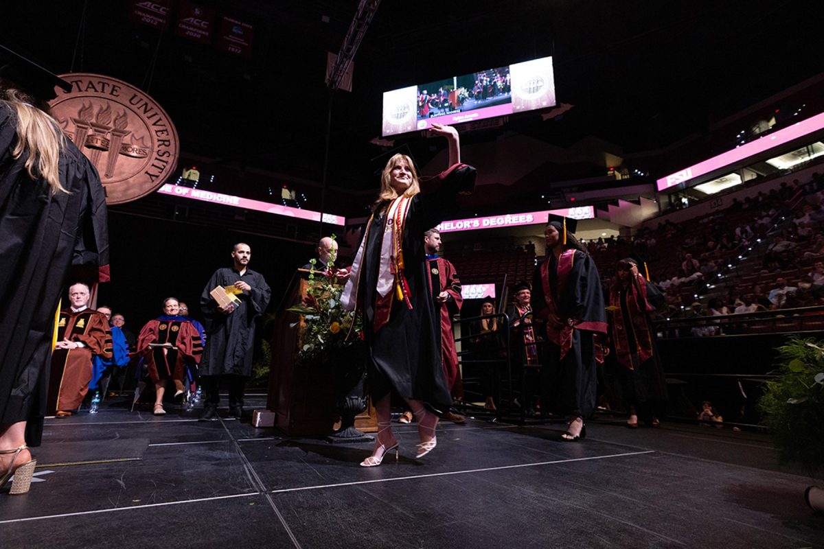 A female graduate wearing a white FSU stole waves to the audience with one hand as she walks across the stage.