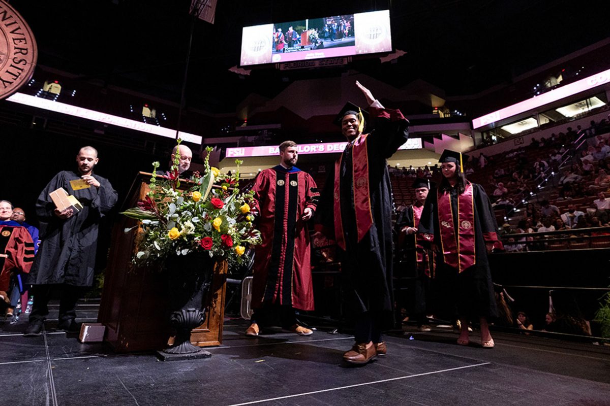 A smiling graduate wearing an FSU stole waves to the audience as he walks across the stage past a floral display.
