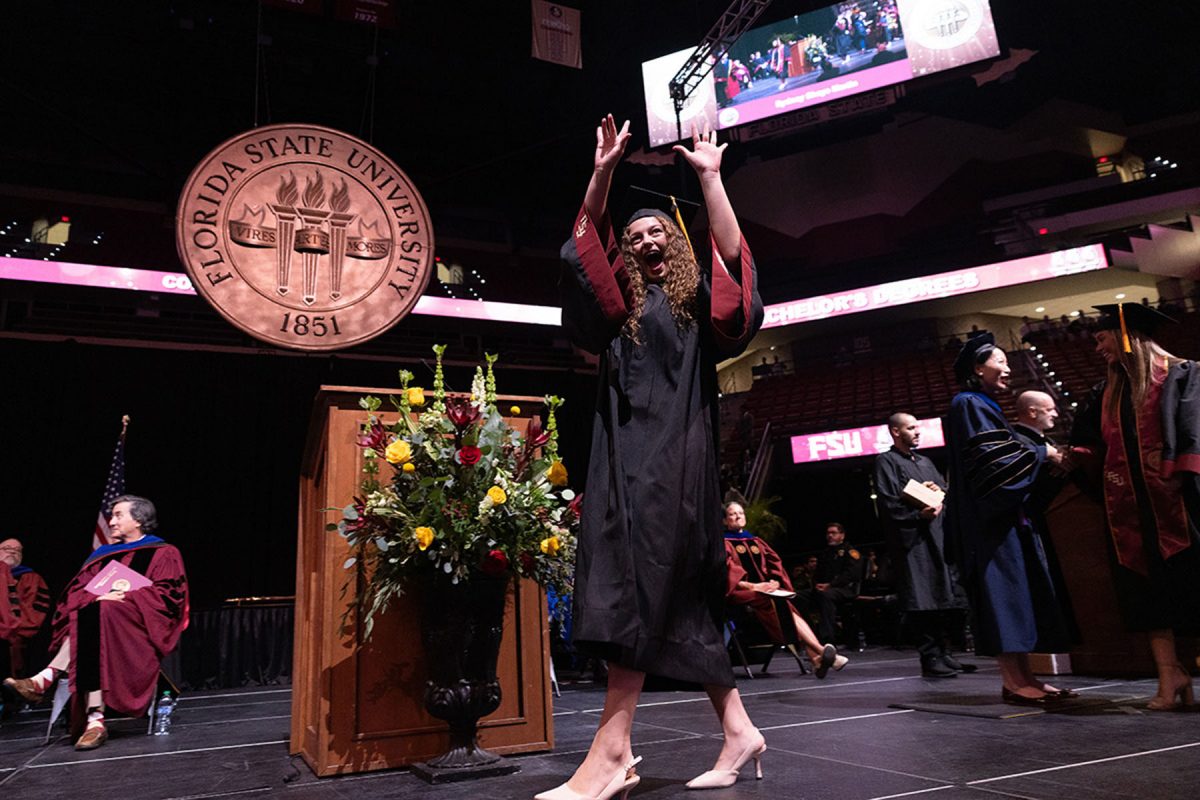 An excited graduate throws both hands up in celebration while walking across the stage beneath the Florida State University seal.