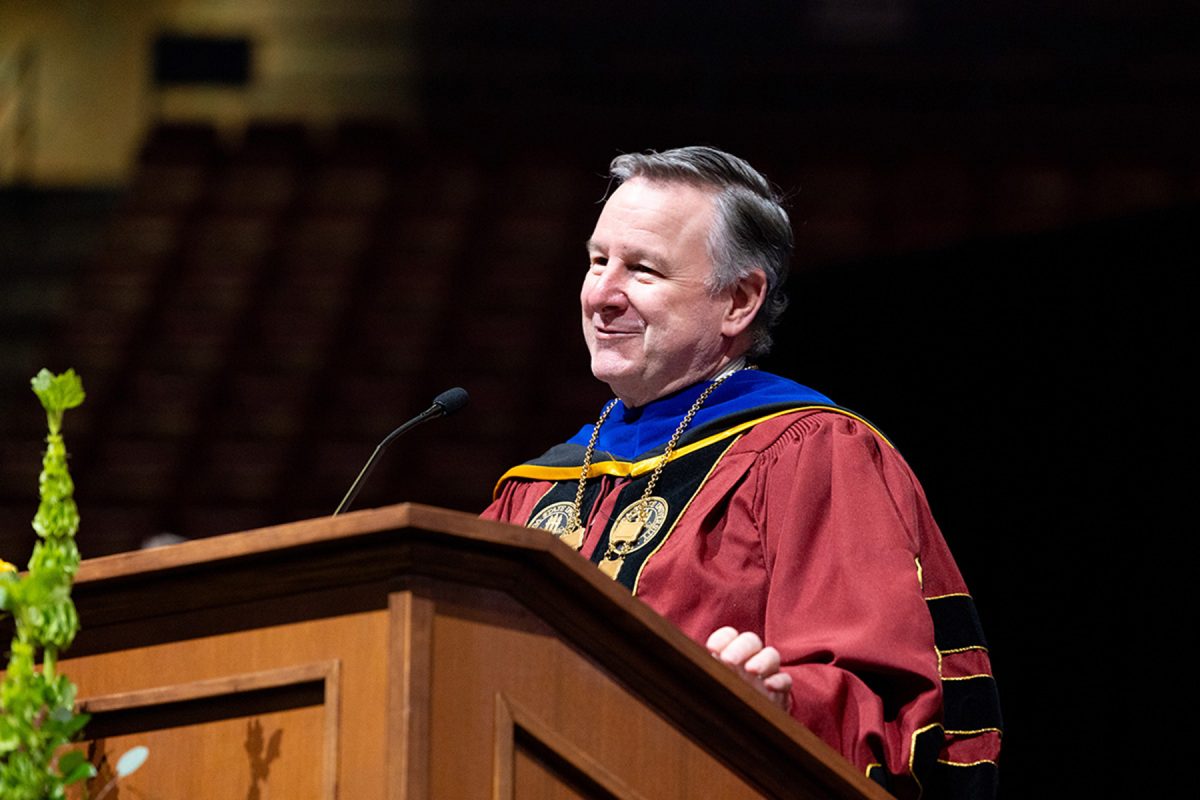 A university official wearing a medallion and academic regalia smiles while standing behind a podium.