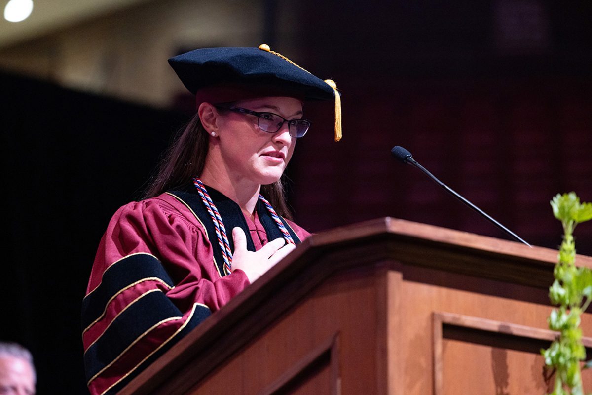A speaker in doctoral regalia holds her hand over her heart while delivering an address from a podium.