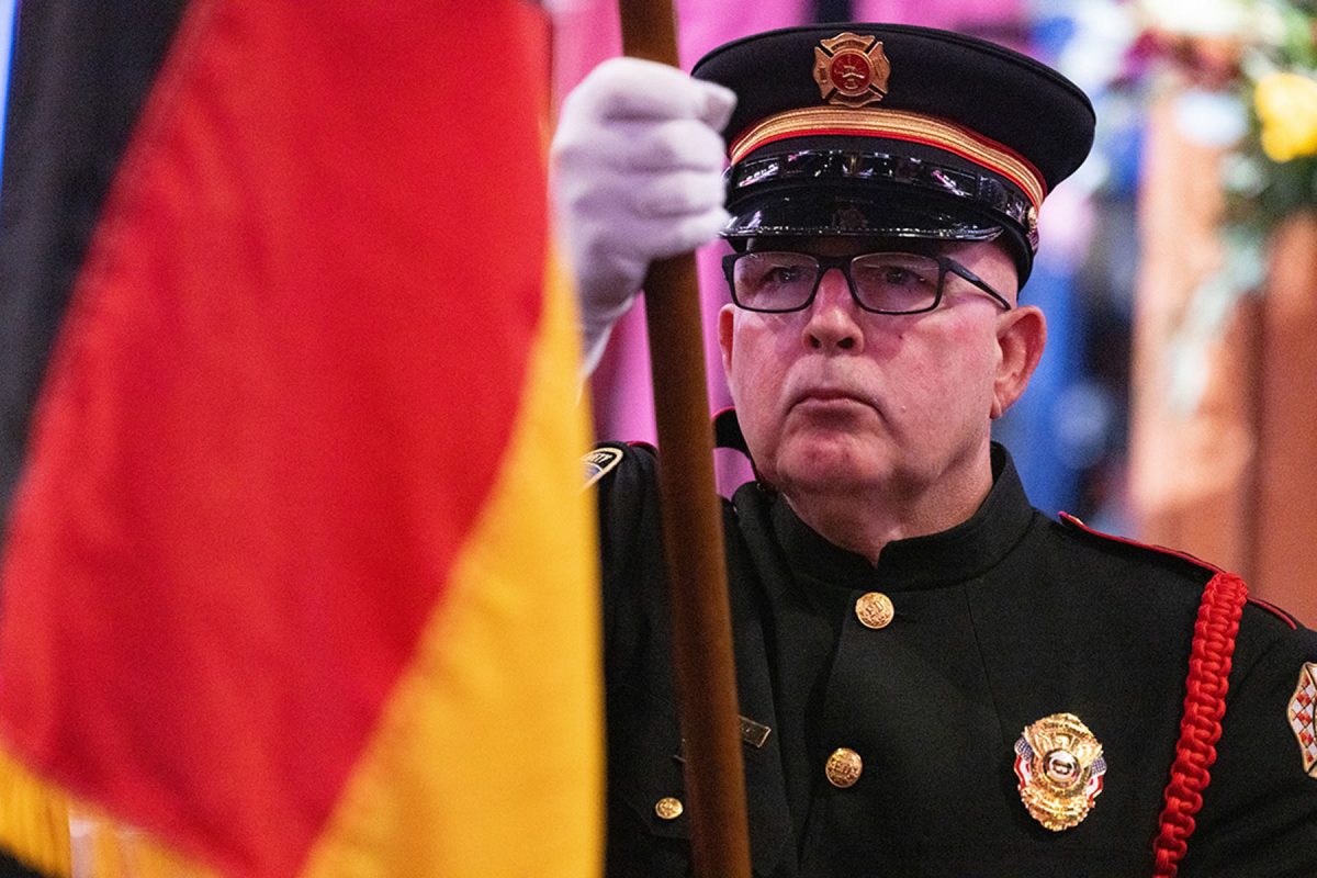 A color guard member in full dress uniform and white gloves holds a flagpole during a ceremonial event.