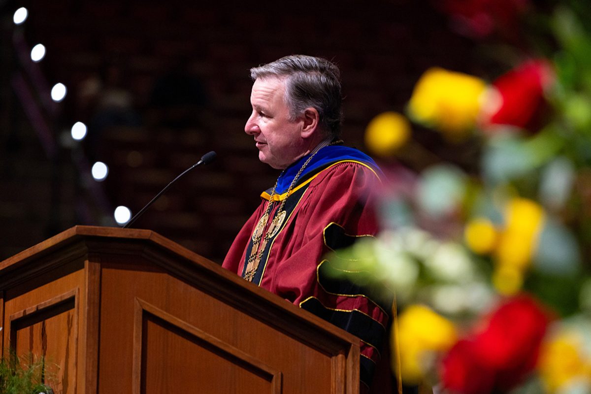 President Richard McCullough in academic regalia speaks into a microphone at a podium decorated with flowers.