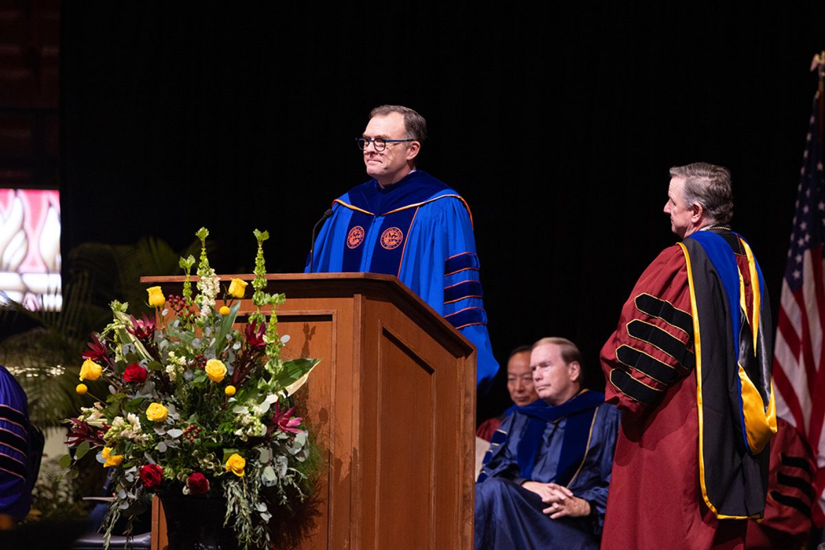 A speaker in a bright blue academic gown stands behind a floral-adorned lectern while addressing the audience.