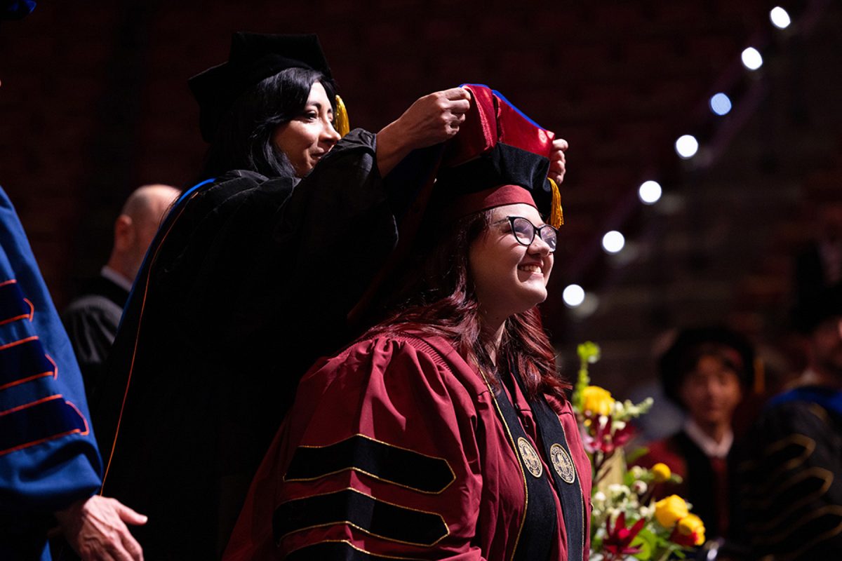 A smiling doctoral graduate wearing glasses looks out into the crowd as her academic hood is draped over her head.