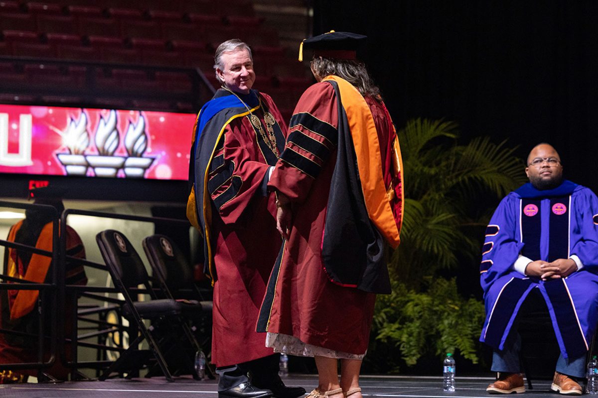 A university leader shakes hands with a newly hooded graduate on stage during a commencement event.