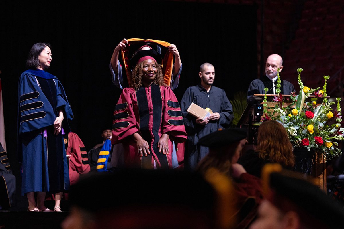 A doctoral candidate smiles as an academic hood is placed over her head during a graduation ceremony.