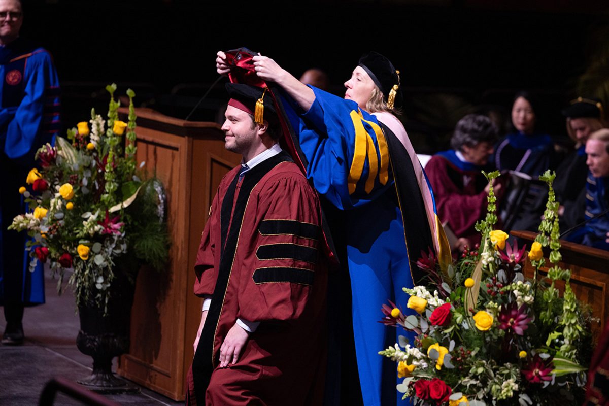 A male doctoral candidate kneels slightly as a faculty member in blue regalia places an academic hood over his head on stage.
