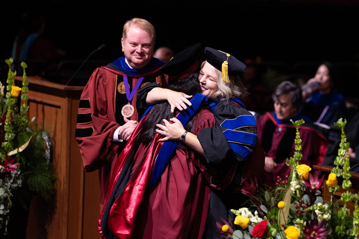 A faculty member warmly embraces a graduate on stage during a graduation ceremony as another official looks on.