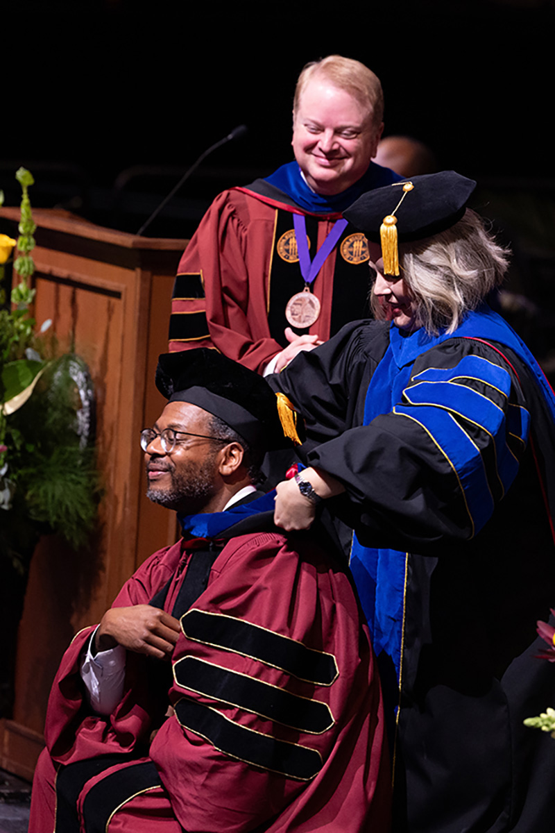 A male graduate wearing glasses and maroon doctoral regalia is being hooded by a faculty member while another administrator watches with a smile.