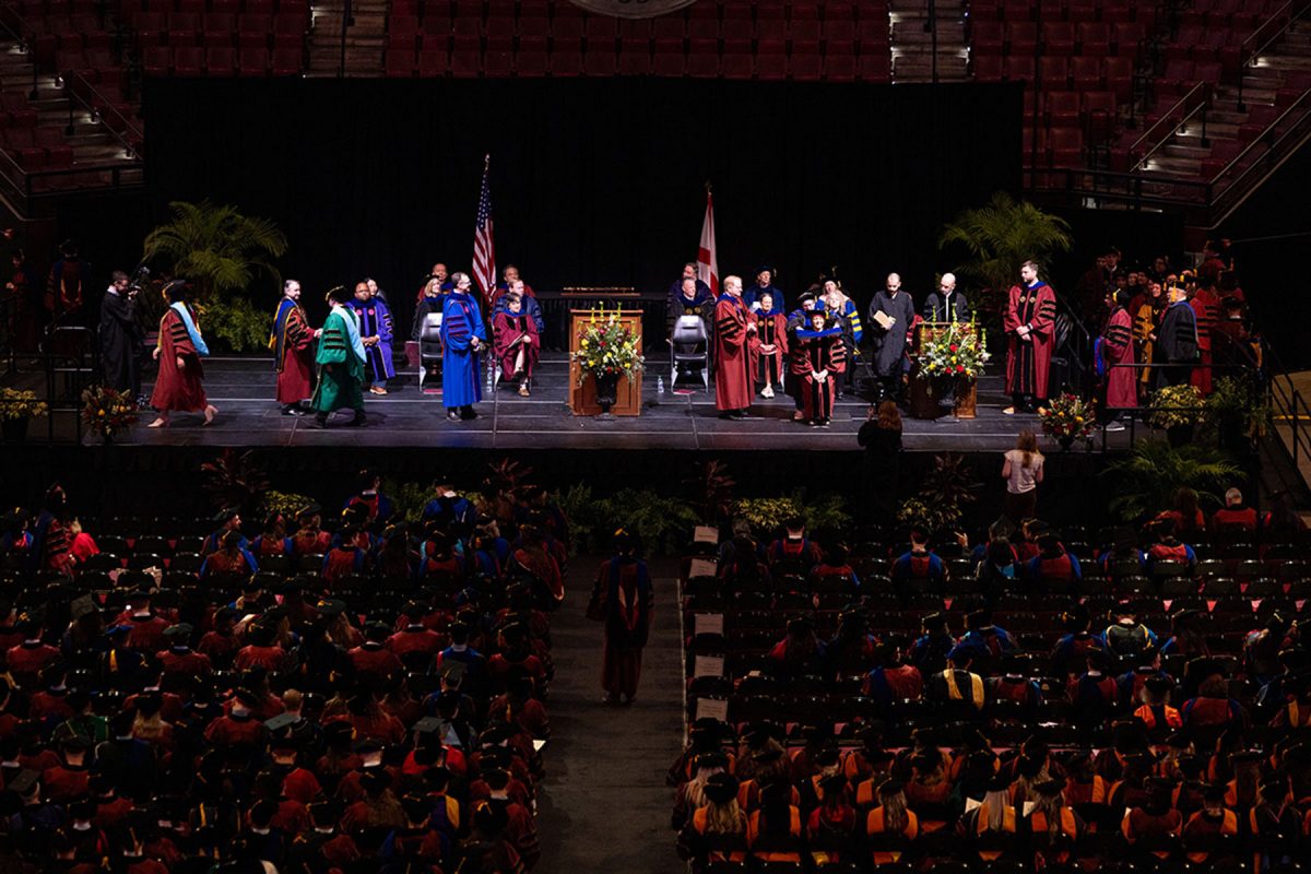 A wide view of a stage and audience seating area inside an arena during a university doctoral hooding ceremony.