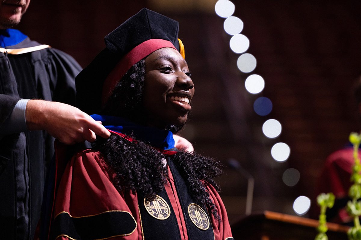 A graduate in doctoral regalia smiles as an academic hood is placed over her shoulders during a commencement ceremony.