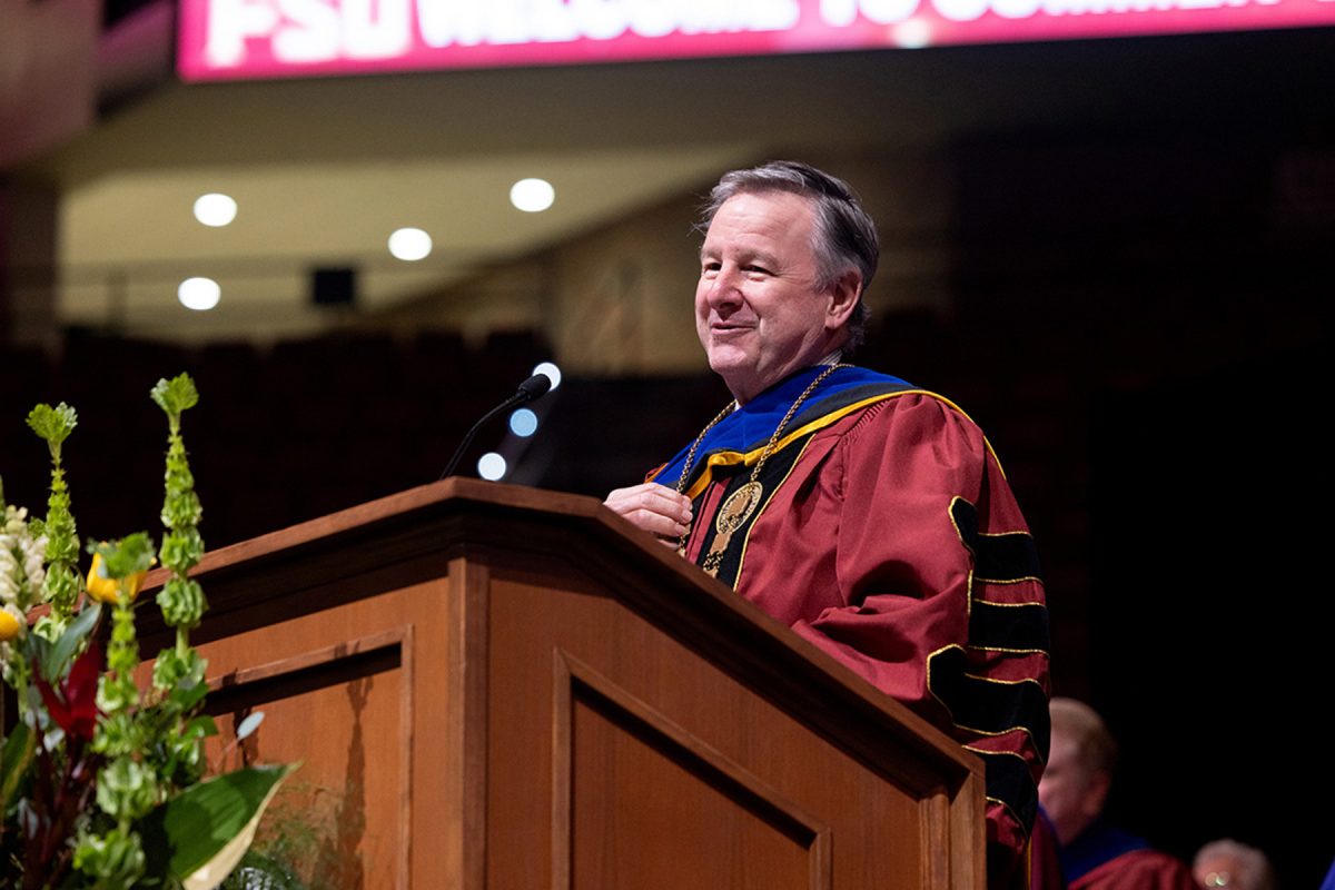 FSU President Richard McCullough, dressed in a maroon gown and ceremonial chain of office, smiles warmly while speaking at a flower-adorned podium.