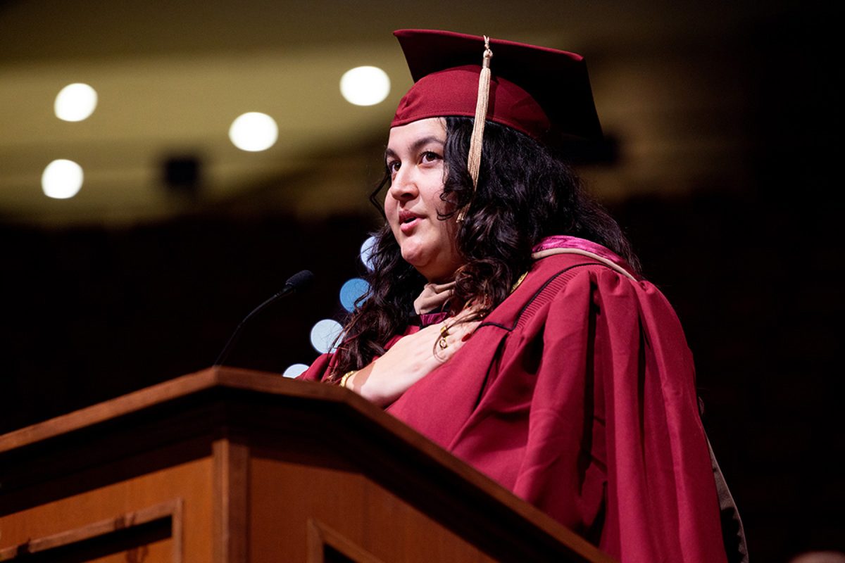 A speaker wearing a maroon cap and gown speaks at a podium with her hand resting over her heart during a commencement ceremony.