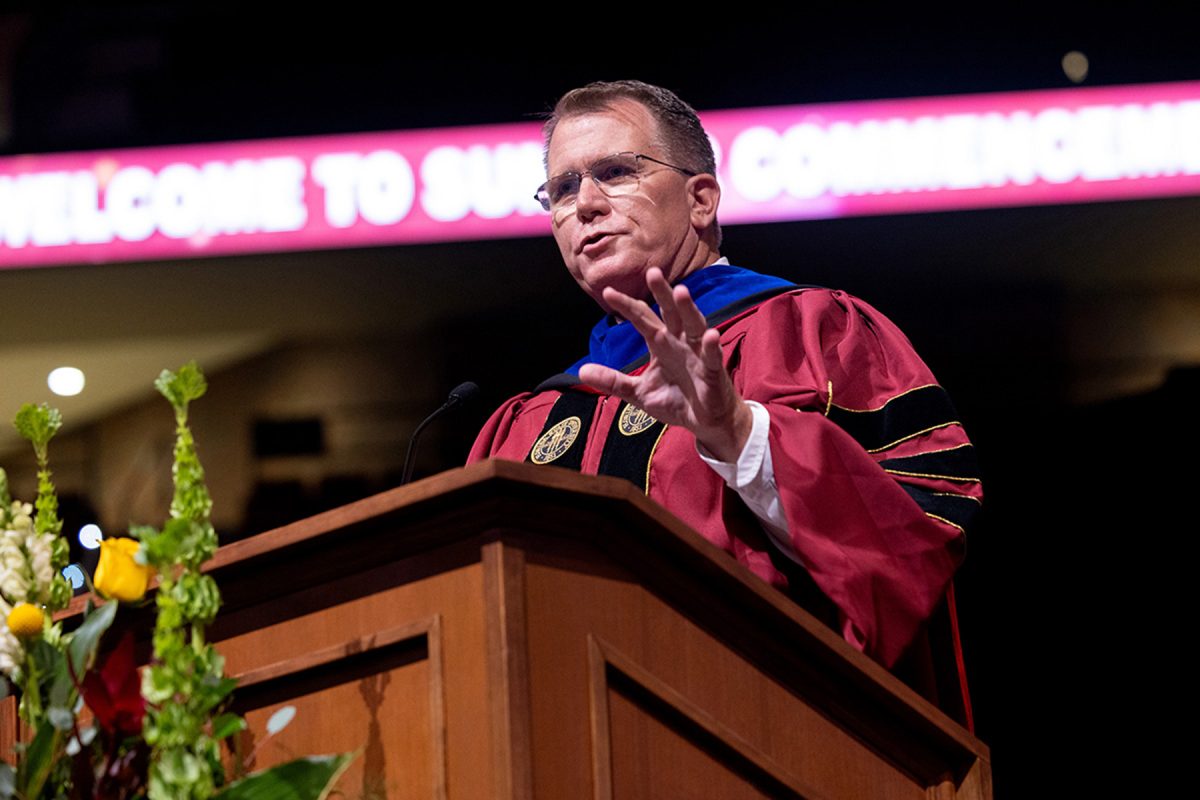 A speaker gestures with his hand while addressing attendees from a wooden podium during a commencement ceremony.