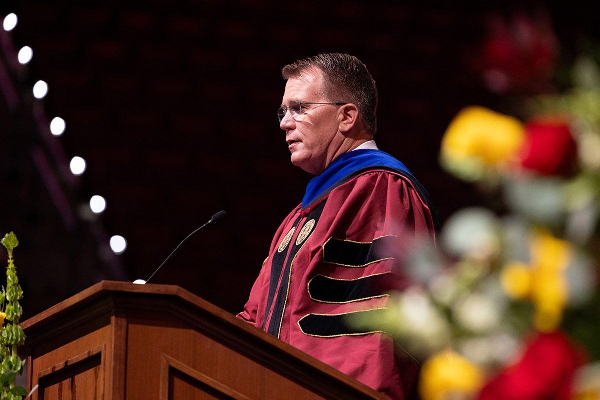 In a side profile shot framed by blurry foreground flowers, a speaker in red academic regalia addresses the audience from behind a wooden podium.