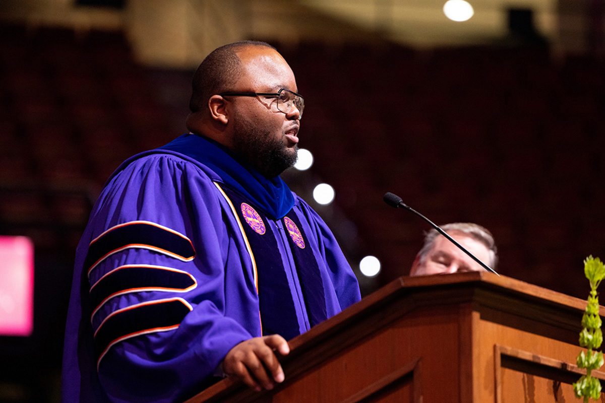 A speaker in purple academic regalia addresses the crowd from a wooden podium during a commencement ceremony.