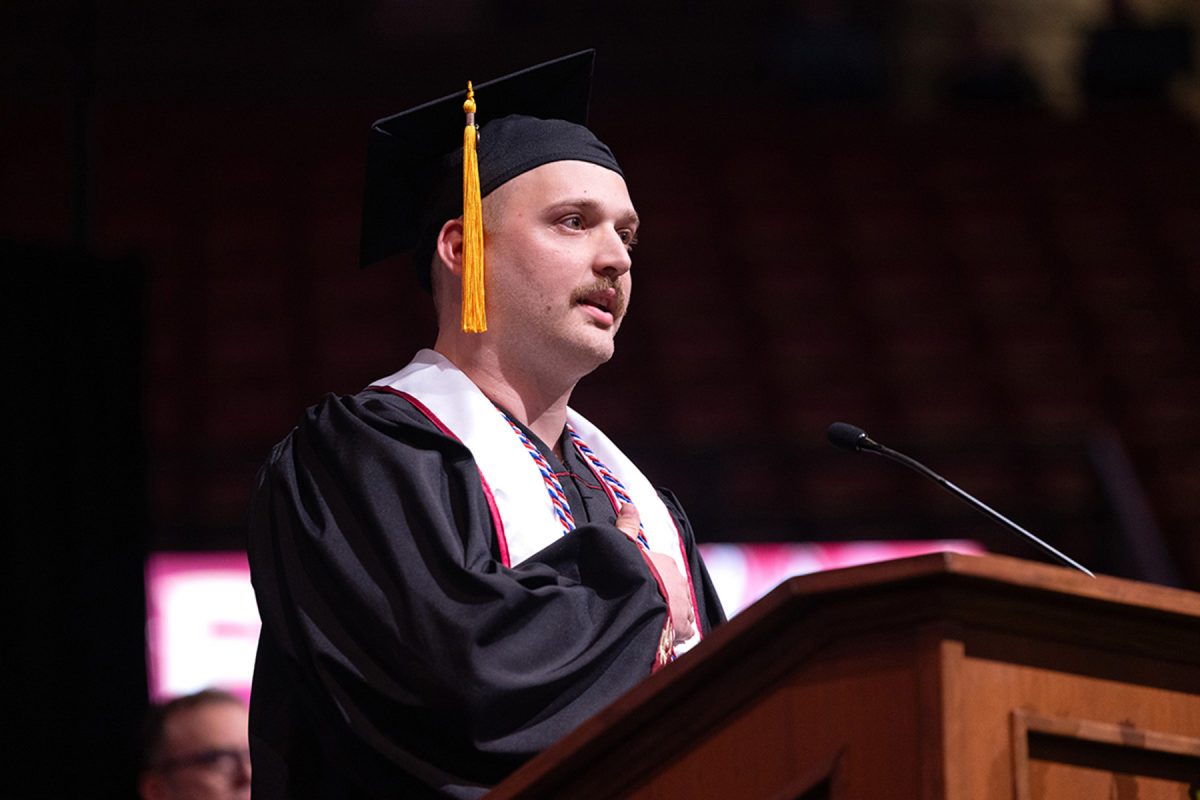 A graduate wearing a black cap, gown, and honor cords speaks at a podium with his hand placed over his heart during the ceremony.