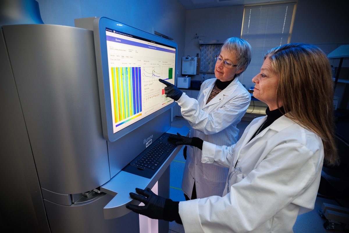 Two female scientists in white lab coats and black gloves review data together on a large touchscreen monitor of an Illumina laboratory sequencing instrument.