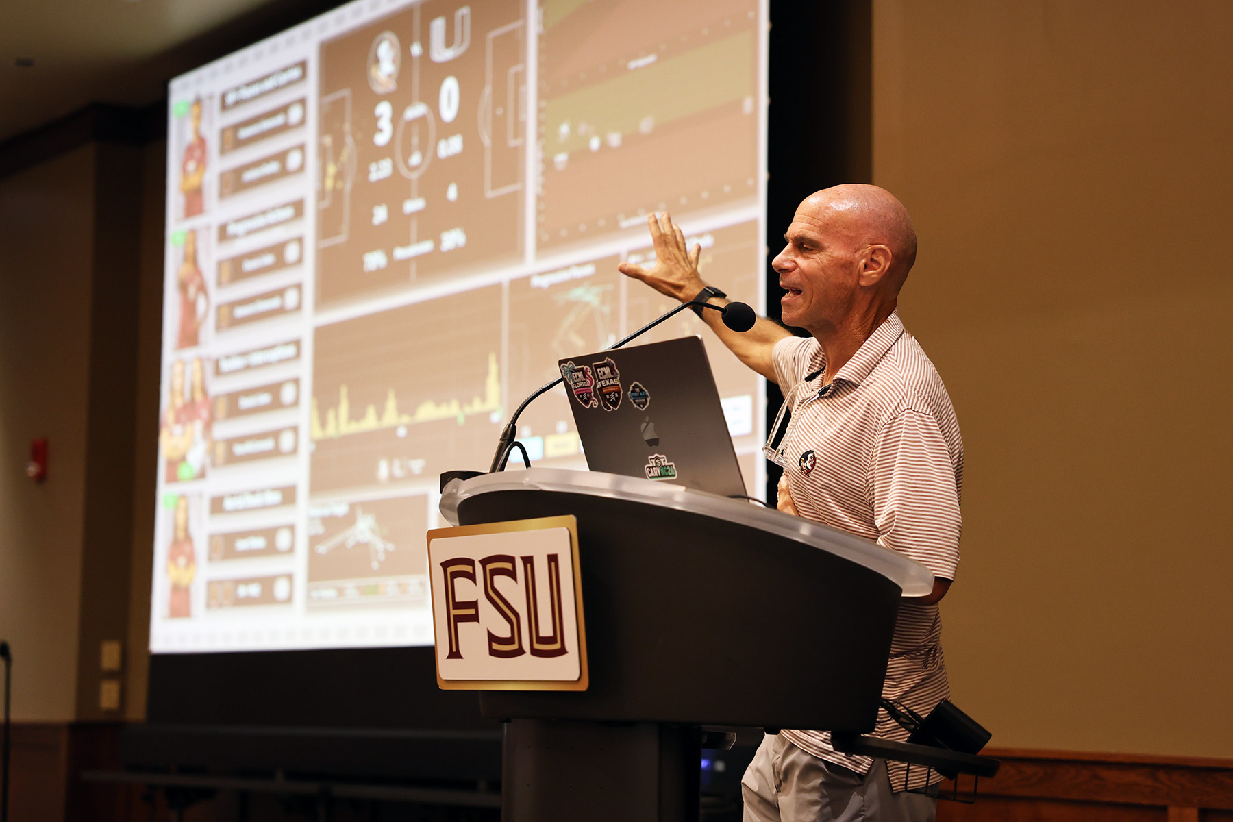 A man stands at a podium branded with the FSU logo, gesturing toward a large presentation screen displaying soccer match analytics.