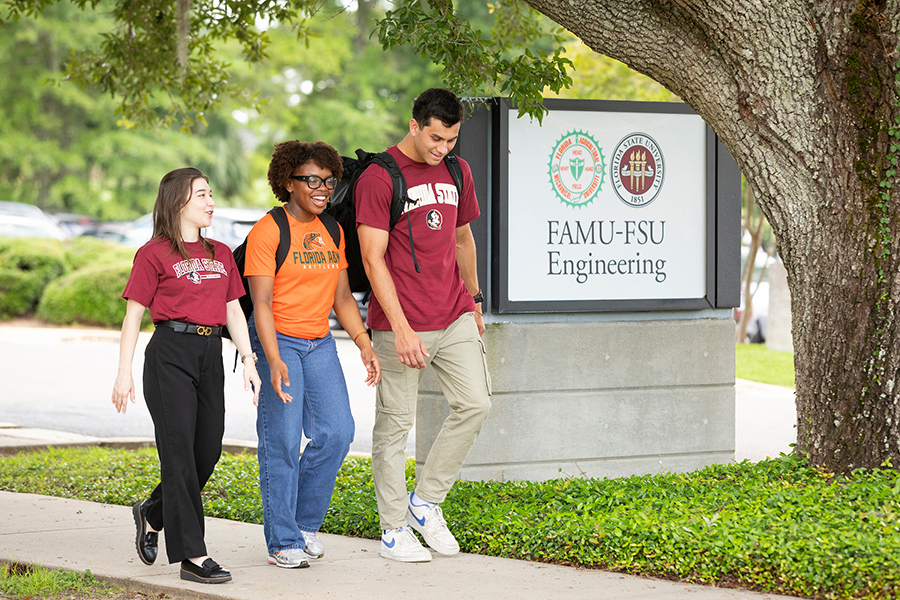 Three students walk together on a campus sidewalk beside a sign reading “FAMU-FSU Engineering.” The students wear university apparel and backpacks while talking beneath a large oak tree.