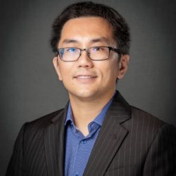 A headshot of a man in a suit wearing glasses on a gray background.