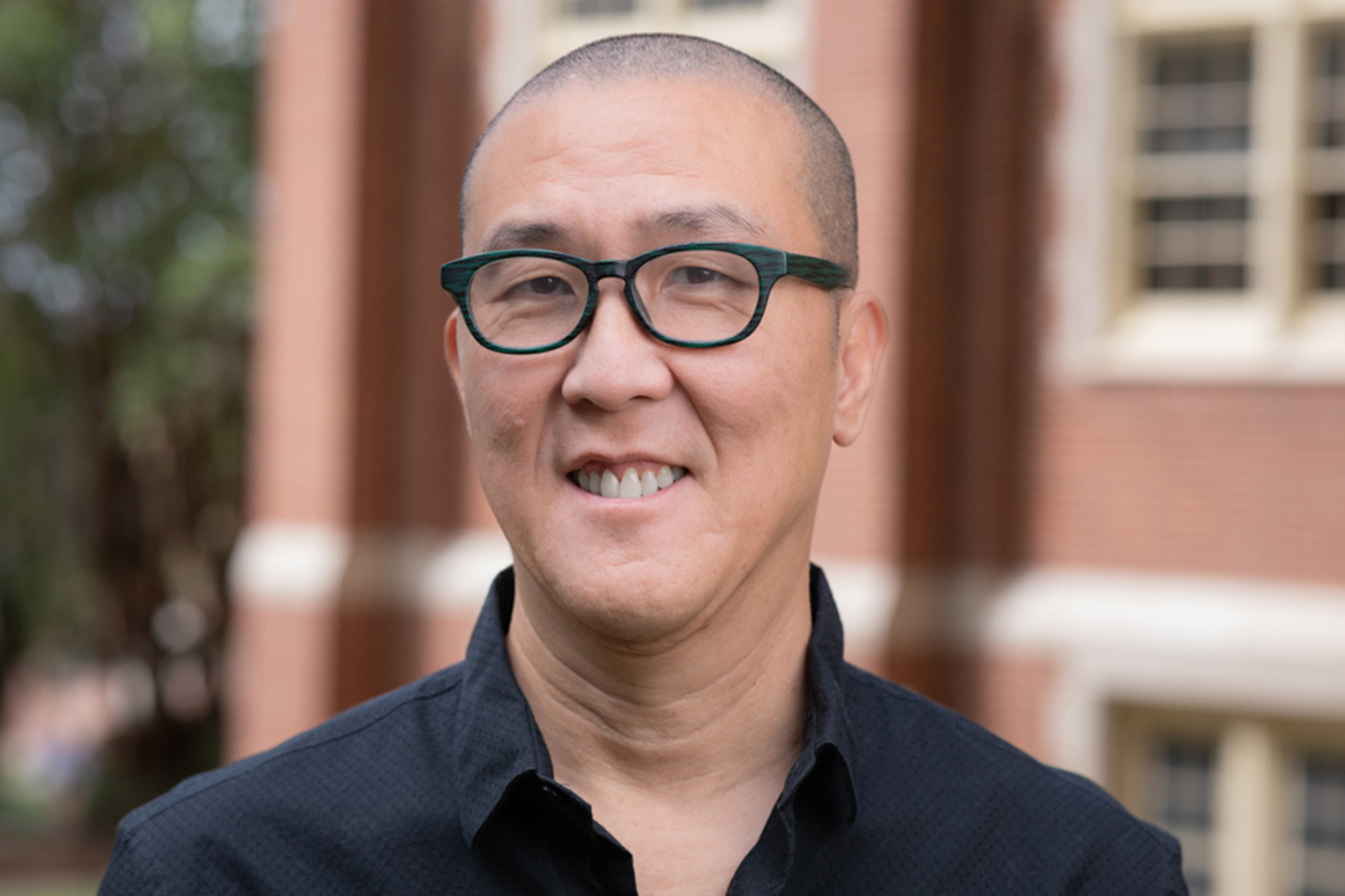 A portrait of a smiling man with a shaved head and glasses, wearing a black button-up shirt in front of a brick building.