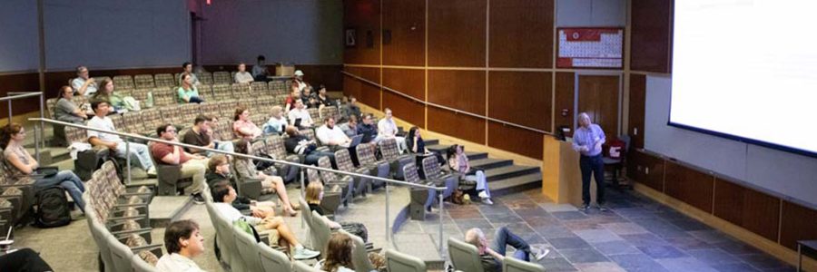 A presenter stands at the front of a lecture hall presenting to a group of seated audience members.