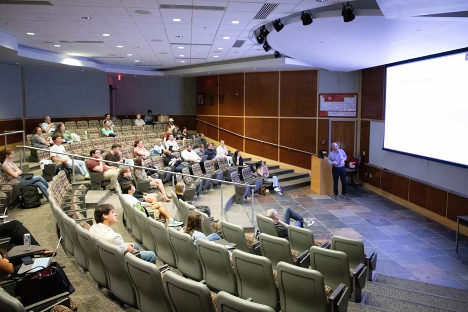A presenter stands at the front of a lecture hall presenting to a group of seated audience members.