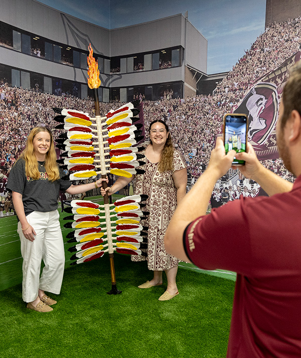 Two women pose together with a flaming spear while someone takes their photo with a smartphone in front of a stadium backdrop.