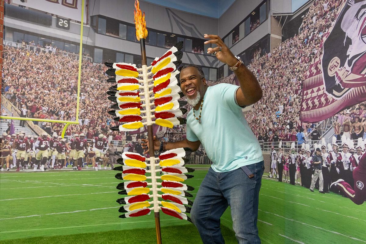 A smiling man poses enthusiastically in front of a stadium backdrop, holding a decorated spear topped with a flaming torch and red, yellow, and black feathers.