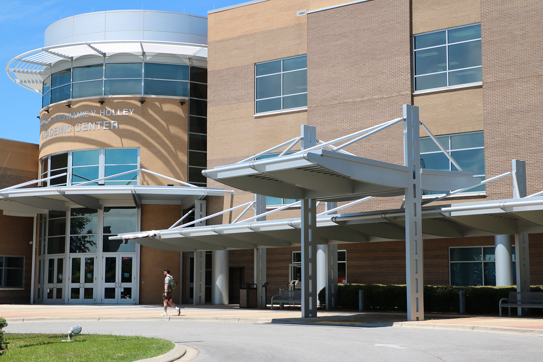 A person with a backpack walks past the entrance of a modern multi-story brick building featuring large glass windows and a prominent covered walkway.