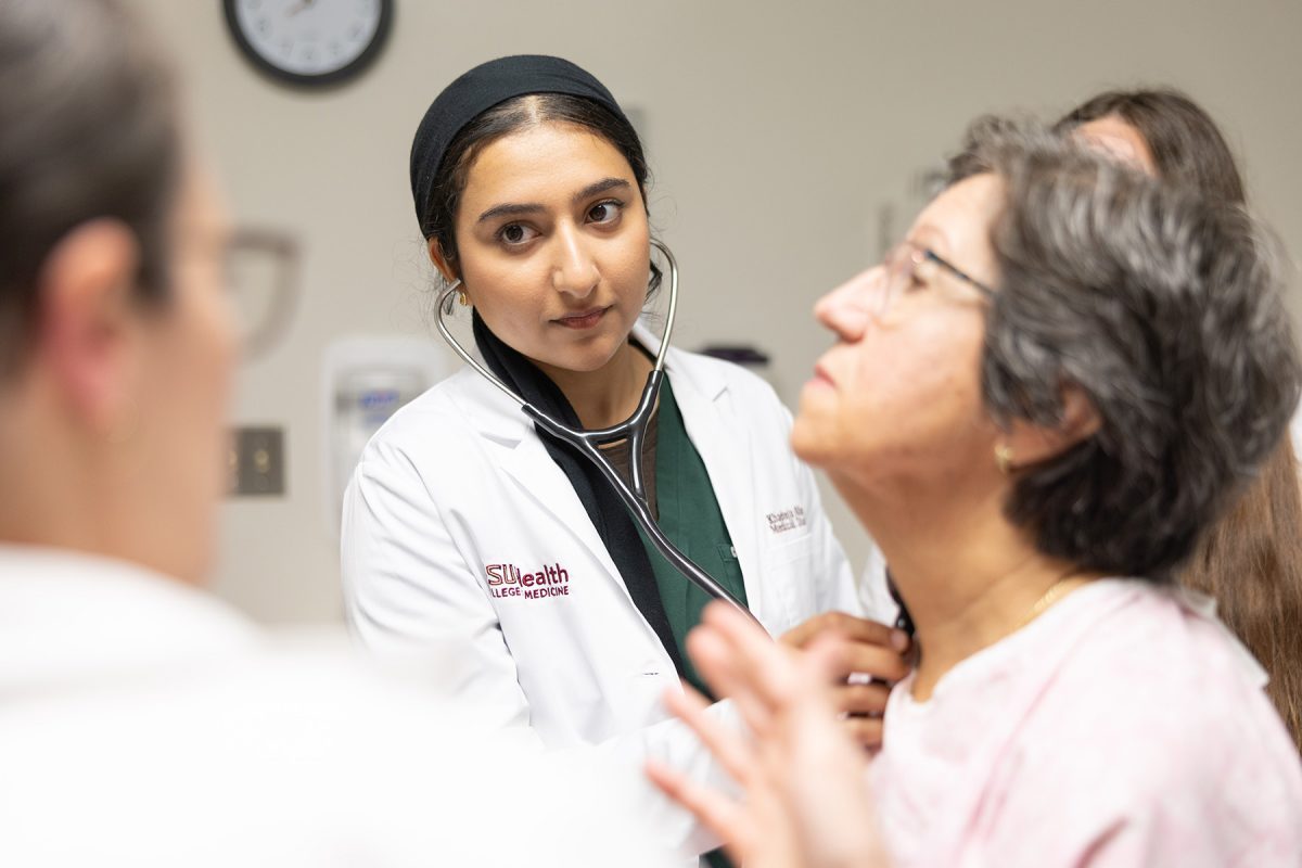 An FSU College of Medicine student wearing a white coat and stethoscope examines a patient while another person observes.