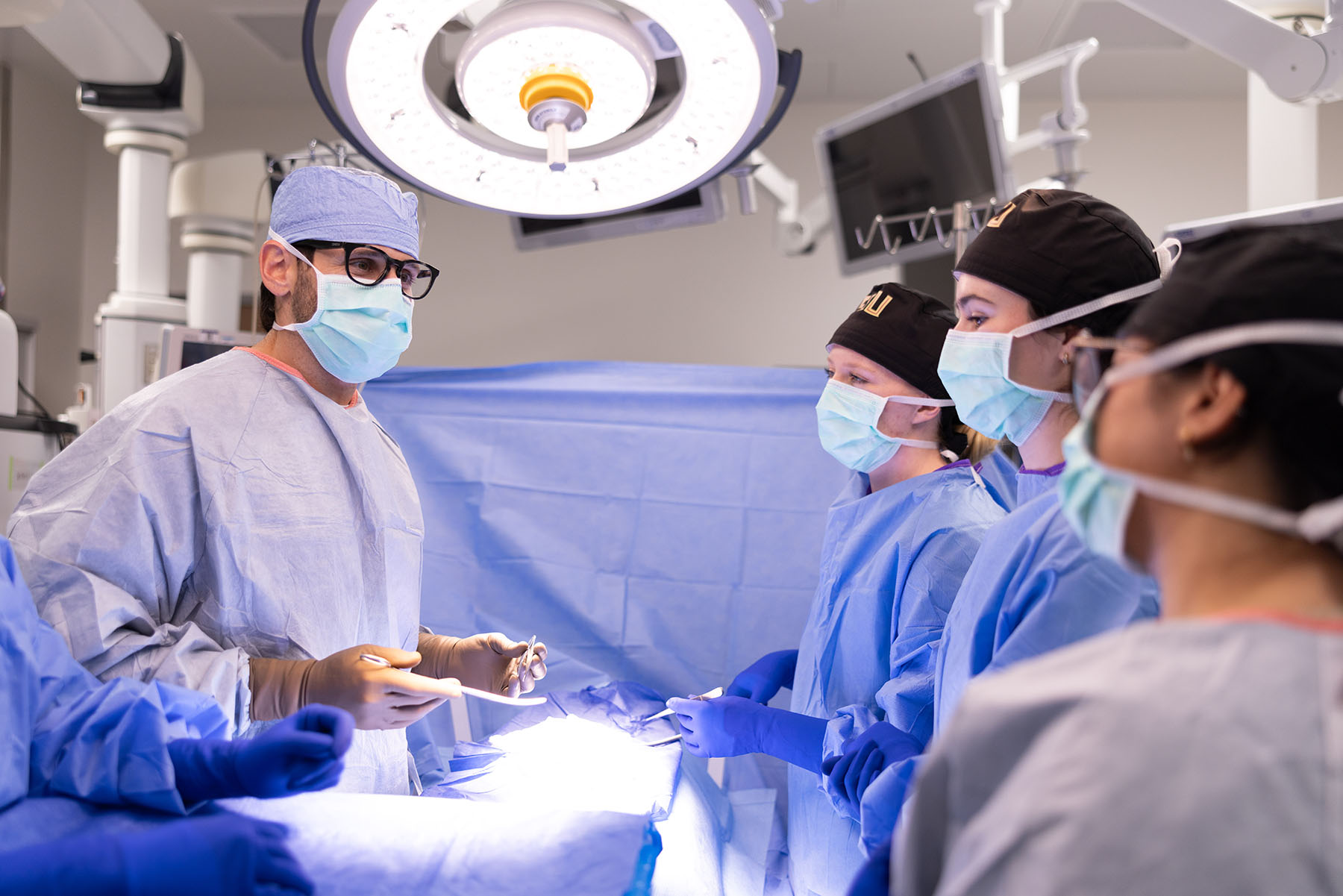 An instructor in surgical attire demonstrates a procedure to a group of masked students wearing surgical gowns in a clinical training room.