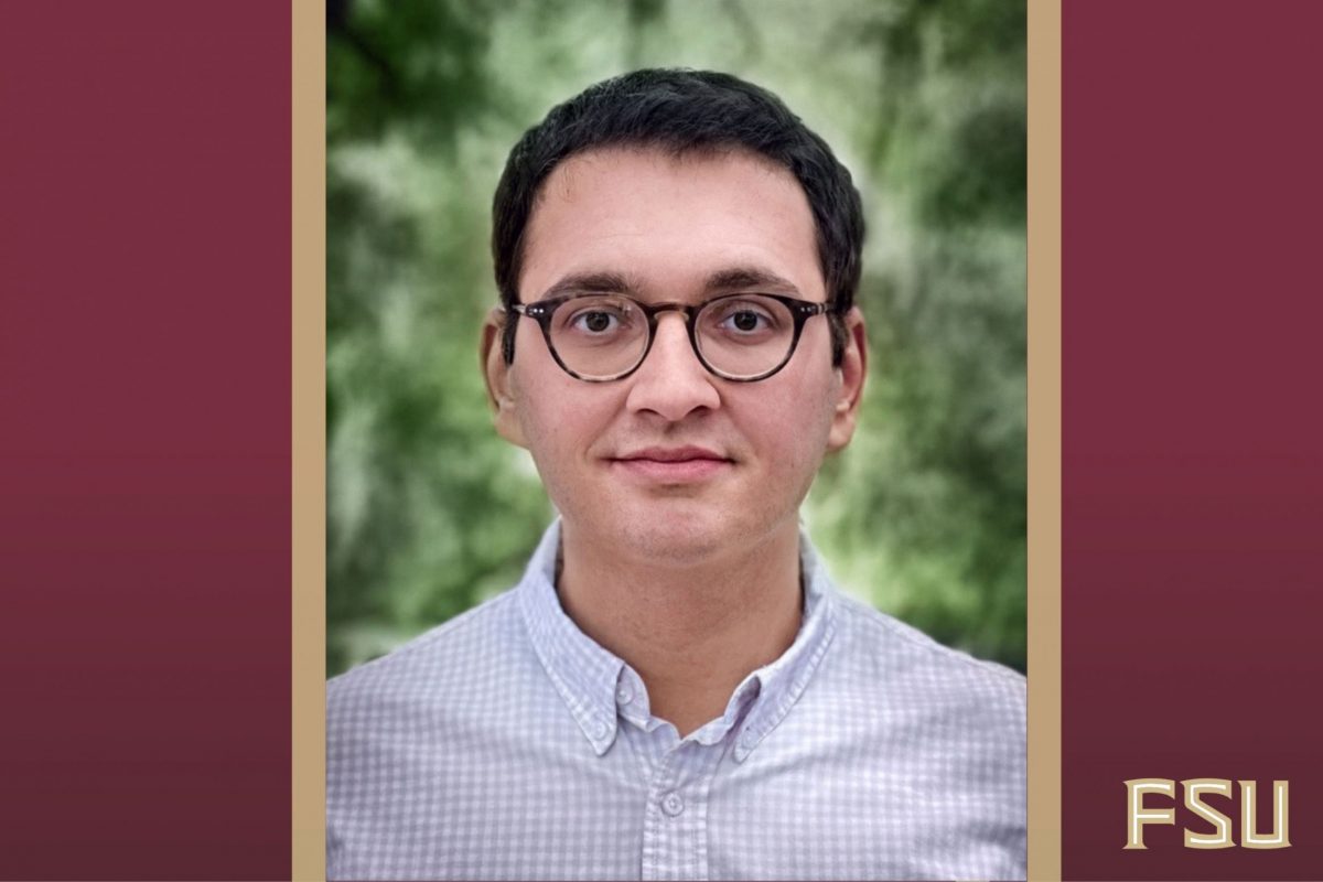 A man in glasses and a blue checkered button down shirt poses for a headshot photo in front of greenery.