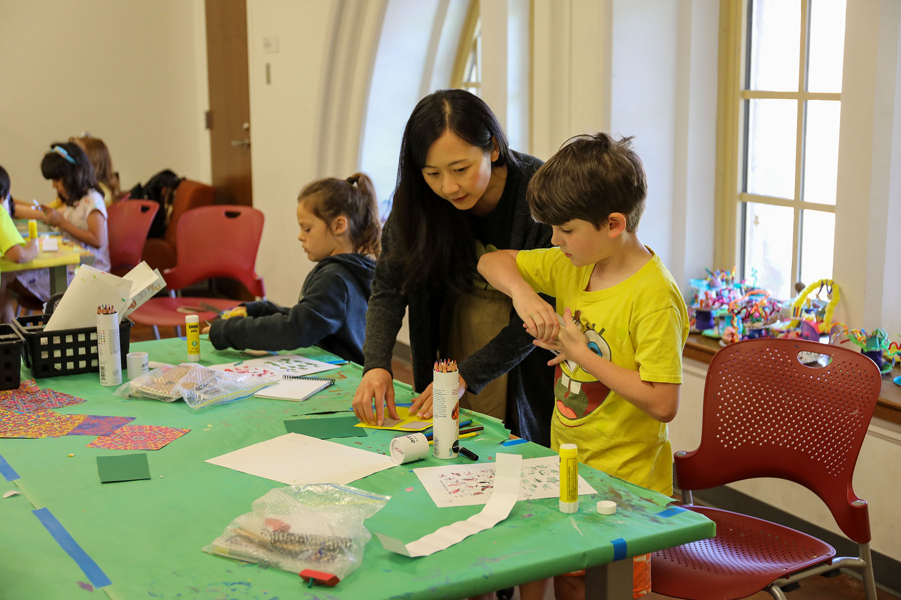 An instructor helps a child with an art project at a table covered with colorful paper, pencils and other art supplies during a K-12 Summer Art Workshop.
