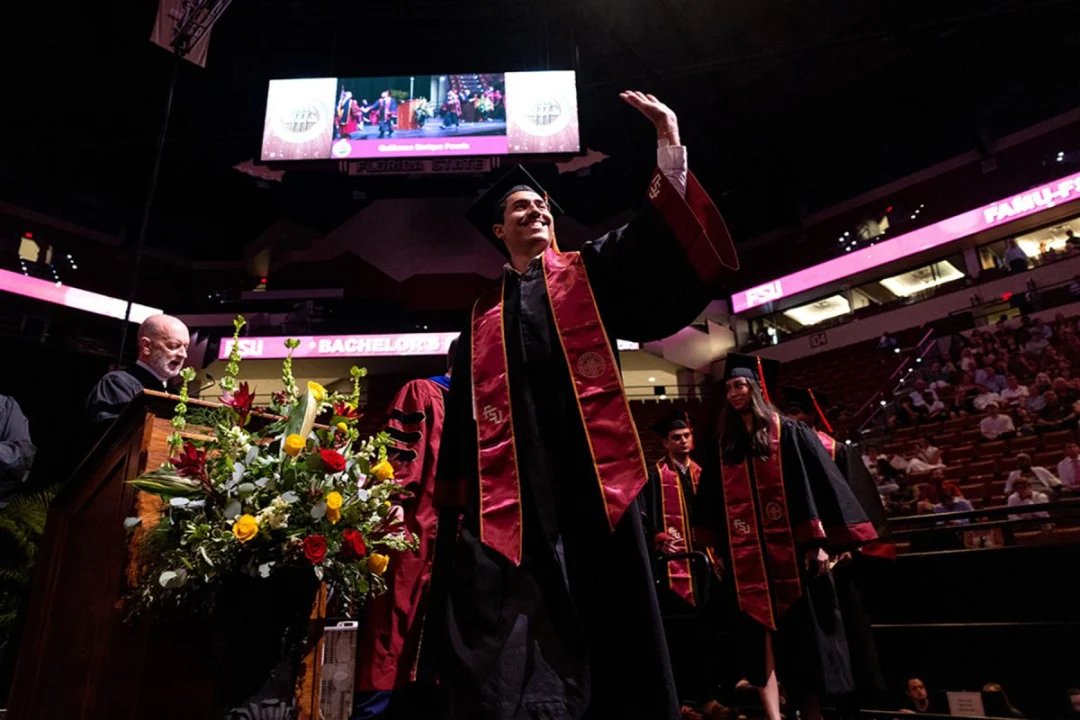 A graduate in a cap and gown waves to the crowd while walking across the stage at a Florida State University commencement ceremony.